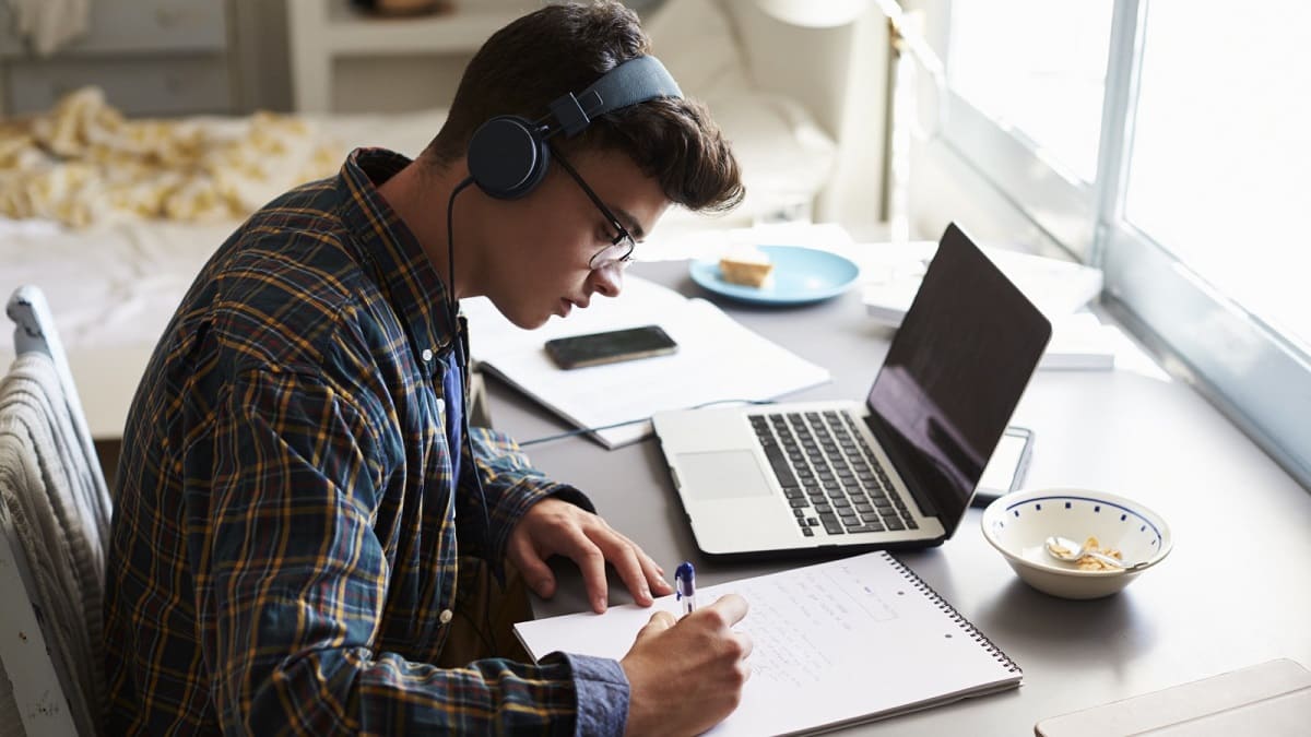 A student wearing headphones writes notes at a desk with a laptop and papers.